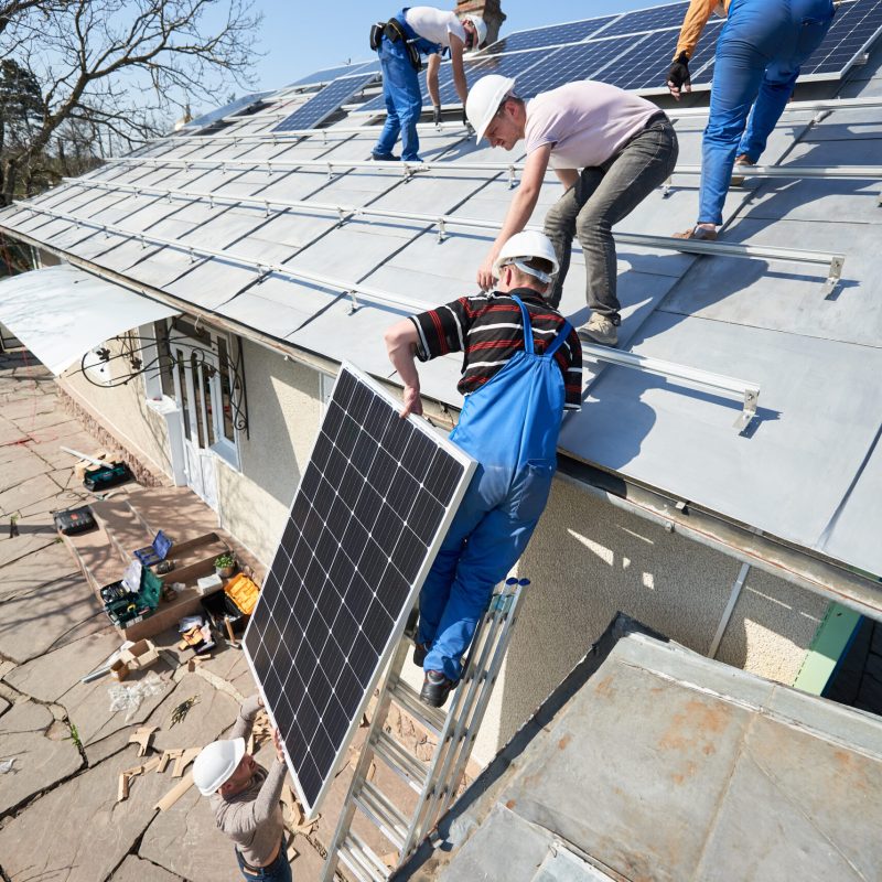 Male team workers installing stand-alone solar photovoltaic panel system. Electricians lifting blue solar module on roof of modern house. Alternative energy ecological concept.